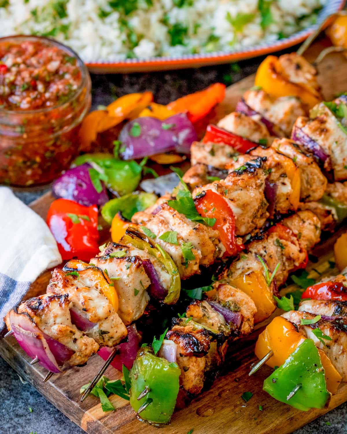 Grilled Mexican chicken skewers with red onion, peppers, and herbs on a wooden board, served with salsa and rice in the background.