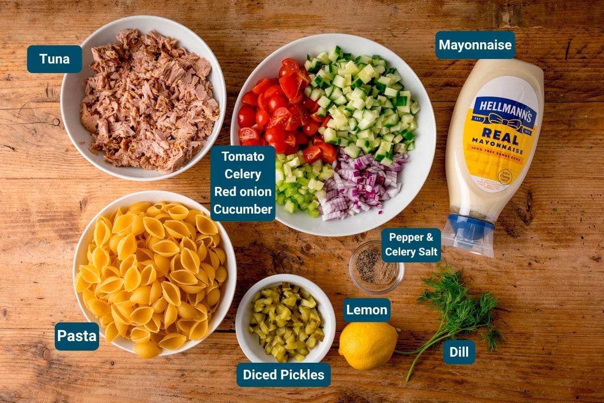 Bowls of pasta shells, tinned tuna, chopped vegetables, diced gherkins, lemon, dill, pepper and celery salt, plus a bottle of mayonnaise are arranged on a wooden surface—everything needed for a classic Tuna Pasta Salad.