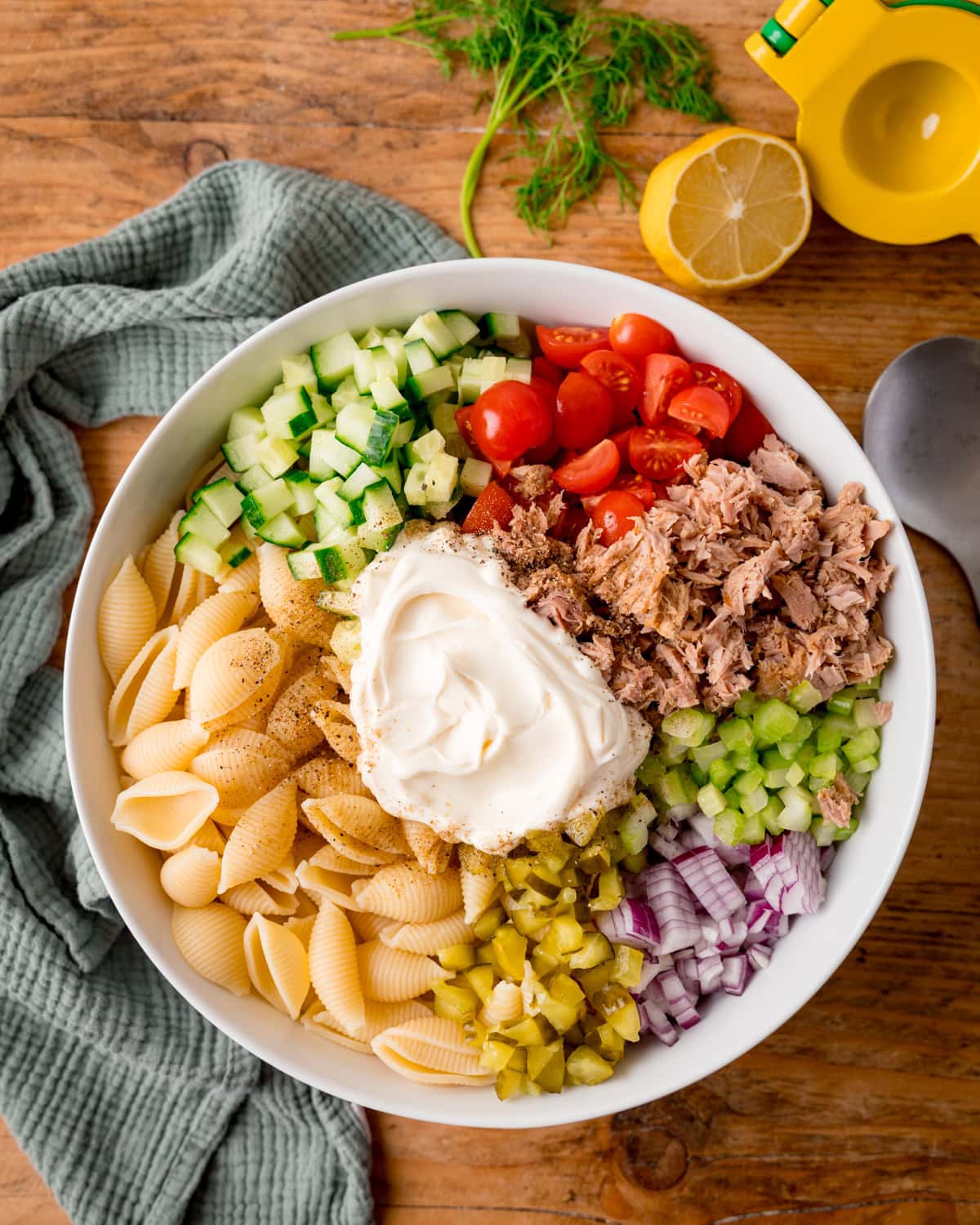 A bowl with cooked pasta shells, chopped cucumber, cherry tomatoes, tuna, celery, red onion, gherkins and a dollop of mayonnaise - ready to be mixed together to make a tuna pasta salad.