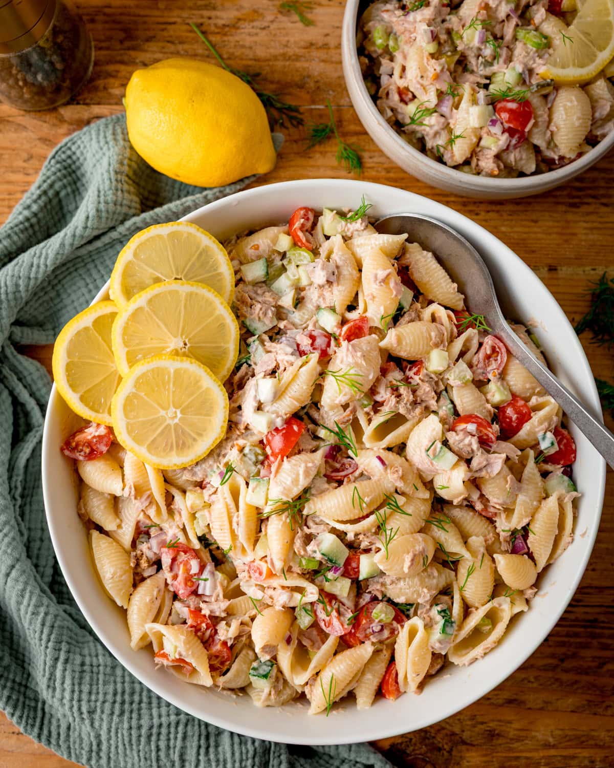 Overhead image of a bowl of Tuna Pasta Salad with shell pasta, tuna, tomatoes, celery, and dill, garnished with lemon slices; a serving spoon and a lemon are close by.