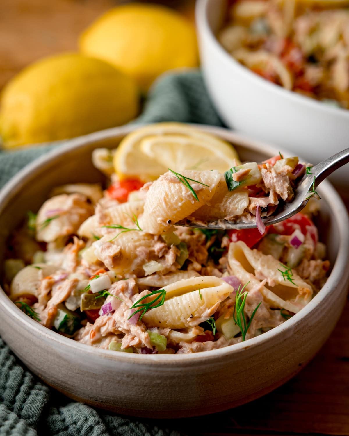 A bowl of Tuna Pasta Salad with pasta shells, tuna, chopped vegetables, herbs, and a lemon wedge, with a fork lifting a bite. Lemons and another bowl of pasta salad are in the background.