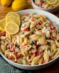 A bowl of Tuna Pasta Salad with pasta shells, tuna, cherry tomatoes, cucumber, fresh dill, and lemon slices, with whole lemons and a smaller bowl of tuna salad in the background.