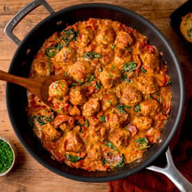 A frying pan filled with Tuscan Chicken Meatballs in creamy tomato sauce with spinach and herbs, on a wooden table with a wooden spoon inside.