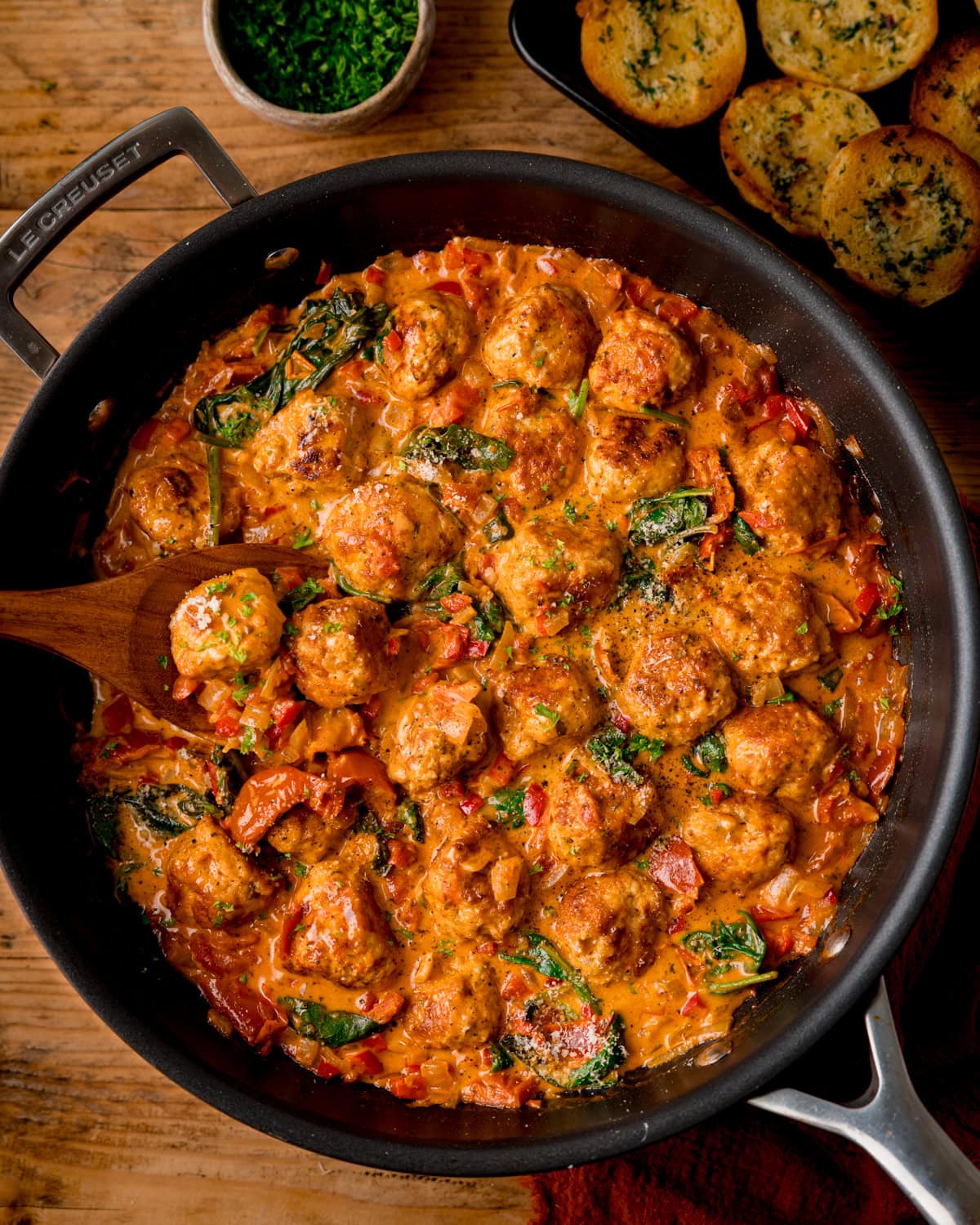A frying pan on a wooden table filled with Tuscan Chicken Meatballs in creamy tomato sauce with spinach and herbs, with garlic bread slices next to the pan.