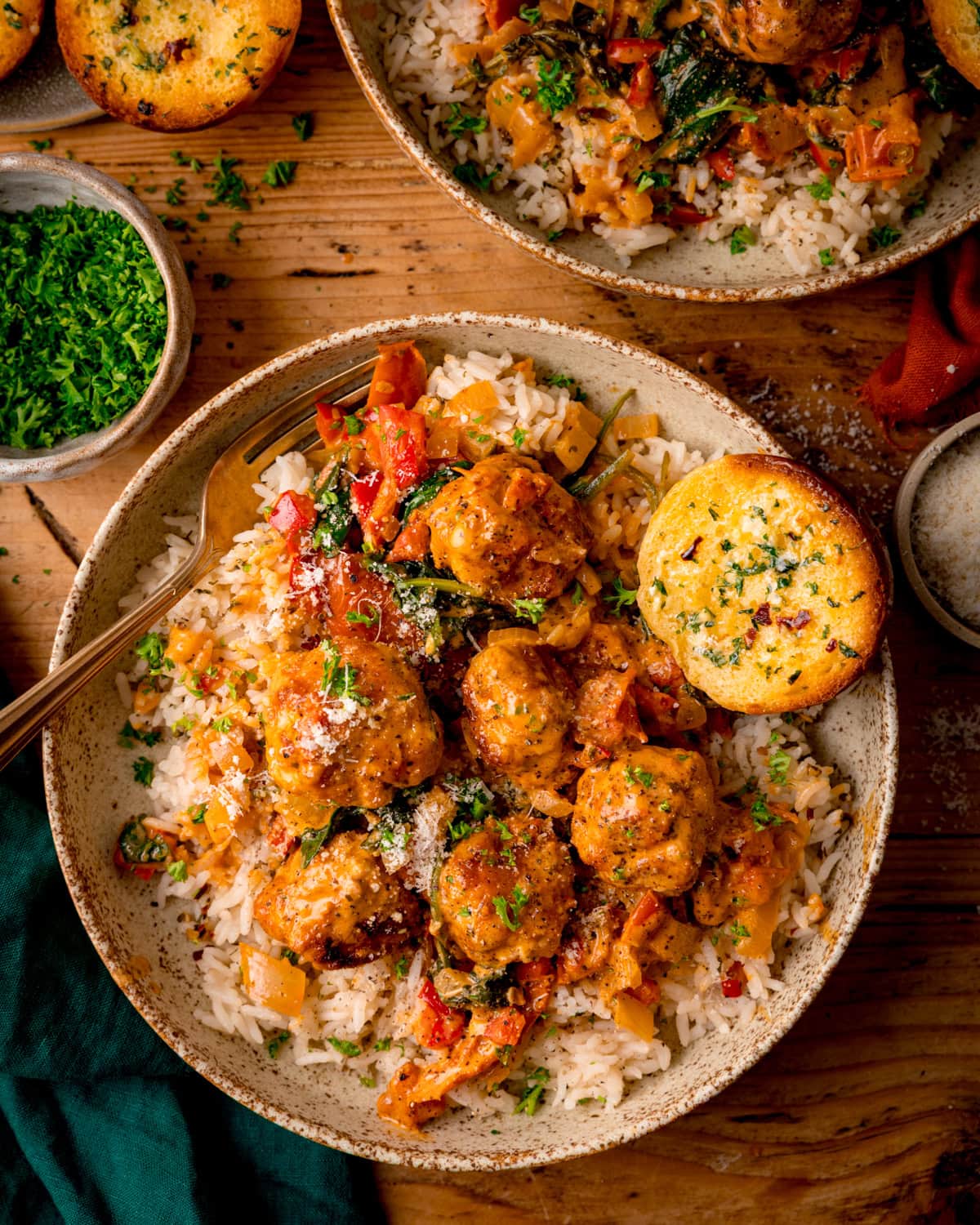 Overhead tall image of a bowl of rice topped with Tuscan Chicken Meatballs, garnished with herbs and grated cheese, served with garlic bread on the side. The bowl is on a wooden table, next to a green napkin, a further bowl of meatballs and chopped parsley.