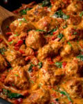 A close up of Tuscan Chicken Meatballs in a creamy tomato sauce with chopped red peppers, spinach, and herbs. A wooden spoon is lifting some of the meatballs out of the pan.