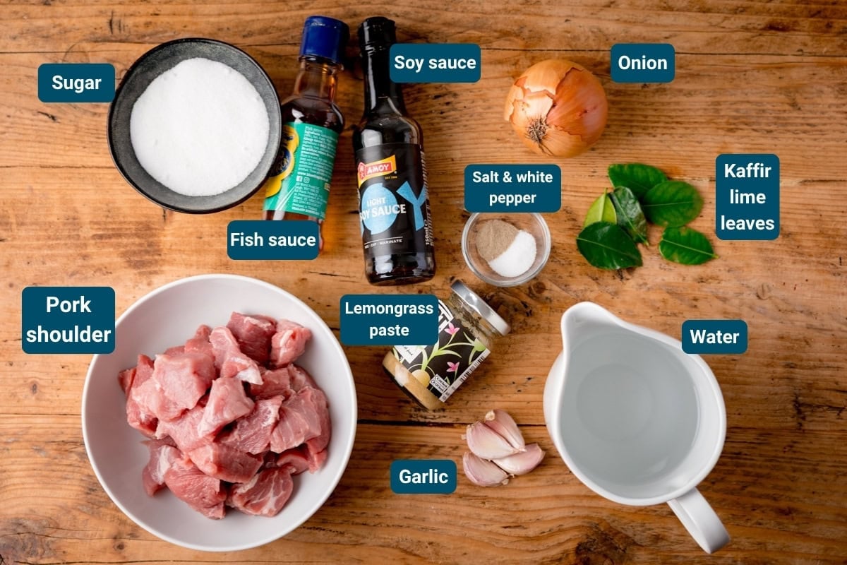 Ingredients for Caramel Pork on a wooden table: pork shoulder, garlic, onion, kaffir lime leaves, lemongrass paste, sugar, fish sauce, soy sauce, pepper and water. All ingredients are labelled.