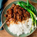 A green plate with white rice, Vietnamese-style caramel pork and grilled spring onions (scallions), with a fork on the side. The plate is on a wooden surface.