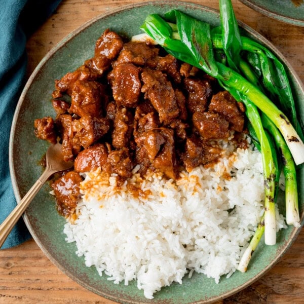 A green plate with white rice, Vietnamese-style caramel pork and grilled spring onions (scallions), with a fork on the side. The plate is on a wooden surface.
