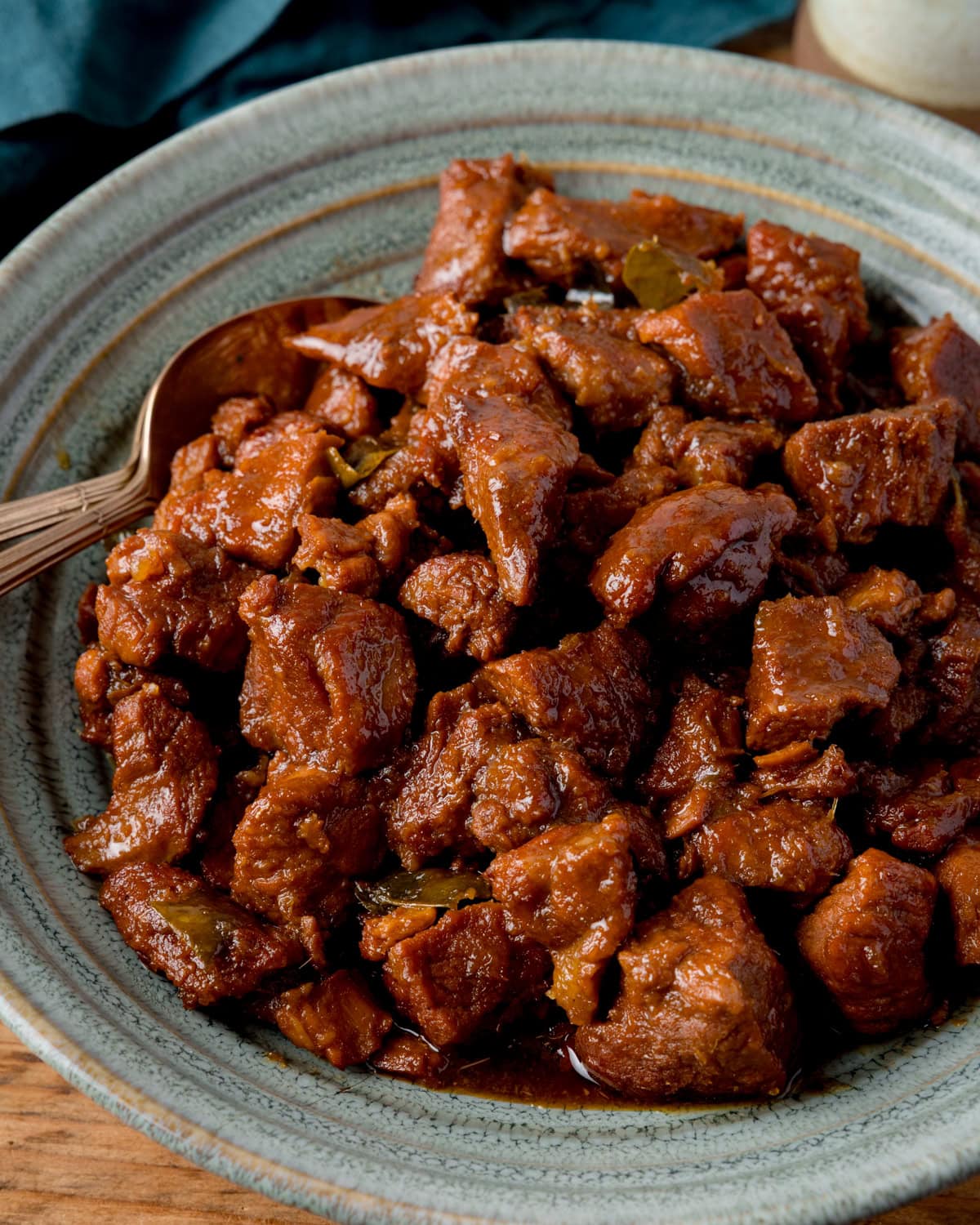 A grey-green bowl filled with saucy, cooked chunks of caramel pork sits a wooden table with two spoons nestled in the pork.