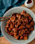 A grey-green bowl filled with saucy, cooked chunks of caramel pork sits a wooden table with two spoons nestled in the pork. A small jug of sauce and a blue-green napkin are nearby.