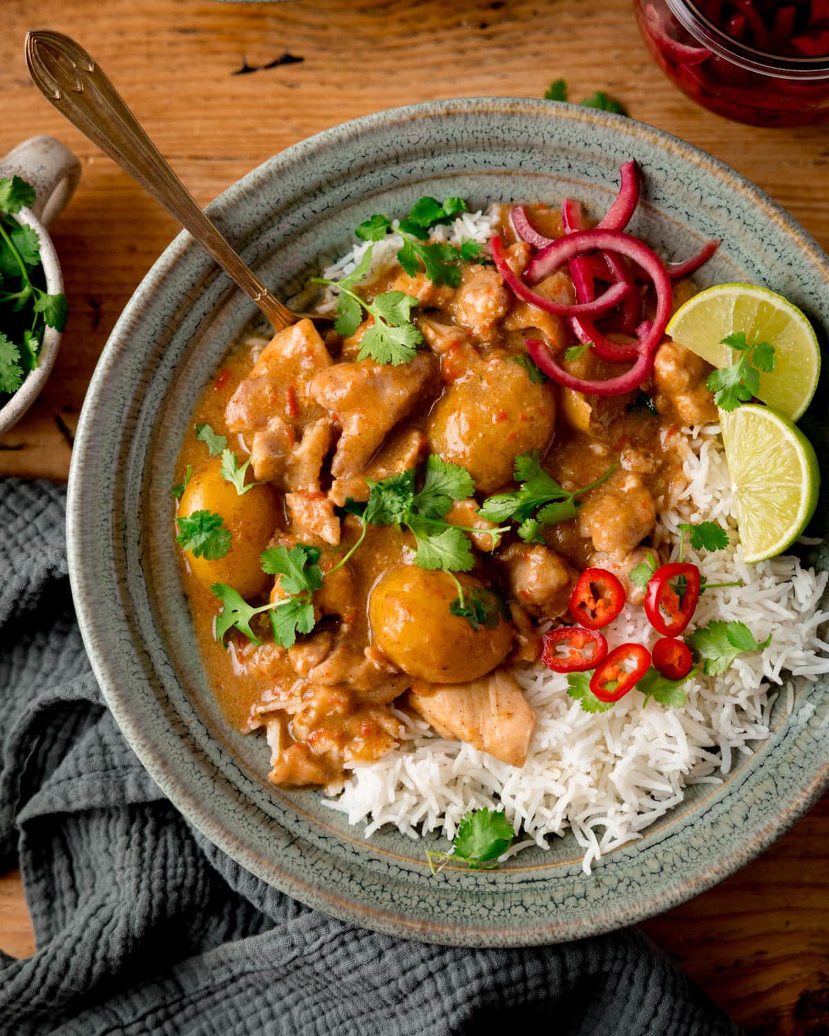 Overhead image of a green bowl filled with Massaman Chicken Curry with potatoes and sauce served over white rice, garnished with lime wedges, red onions, sliced red chillies, and coriander. The bowl is on a wooden surface and there is a sage green napkin next to it.
