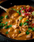 Close up of a pan of Massaman Chicken Curry with baby potatoes, topped with fresh coriander and pickled red onions, with a wooden spoon resting in the pan.