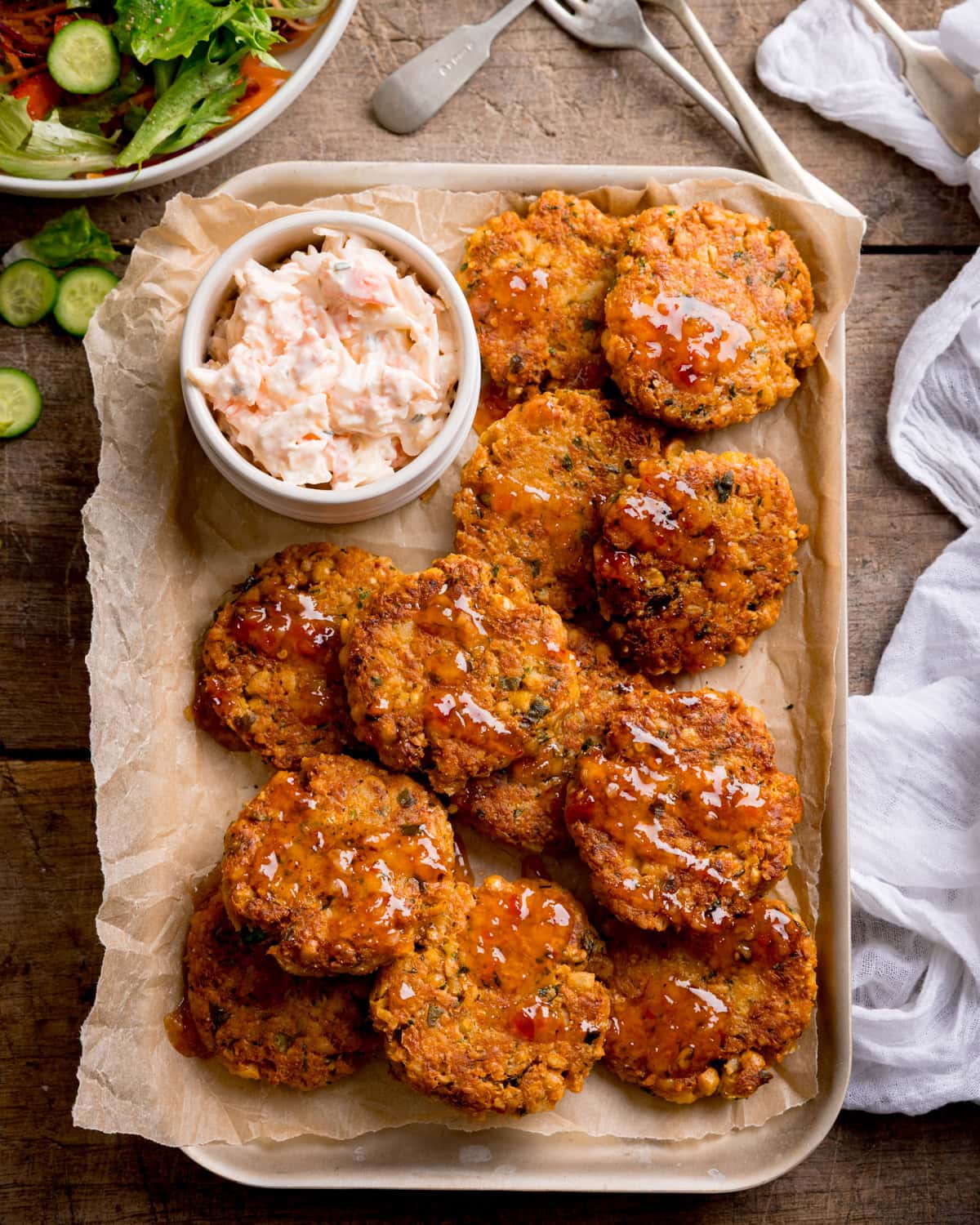A tray of chickpea patties topped with sauce, served with a bowl of coleslaw on greaseproof paper. A green salad is partially visible in the background.