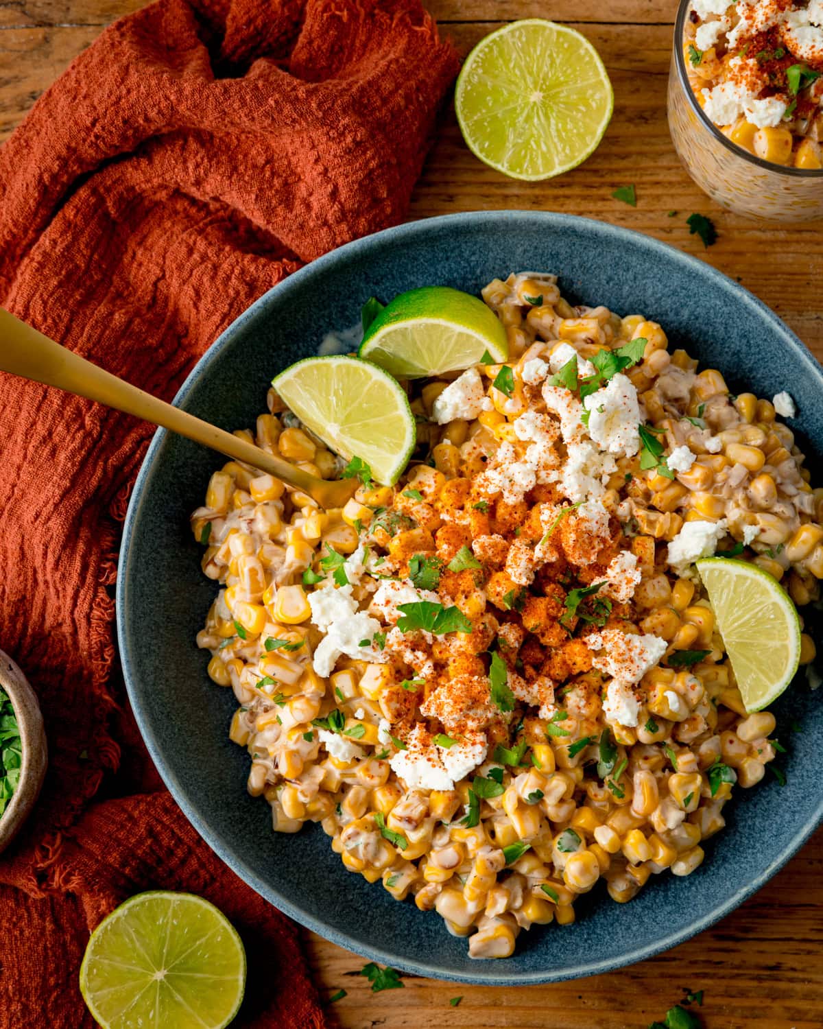Overhead image of a blue bowl filled with Mexican street corn salad, also called Corn Esquites, topped with crumbled cheese, Tajin powder, and lime wedges, with a gold spoon in the corn. The bowl is on a wooden surface, next to a terracotta napkin and lime pieces.