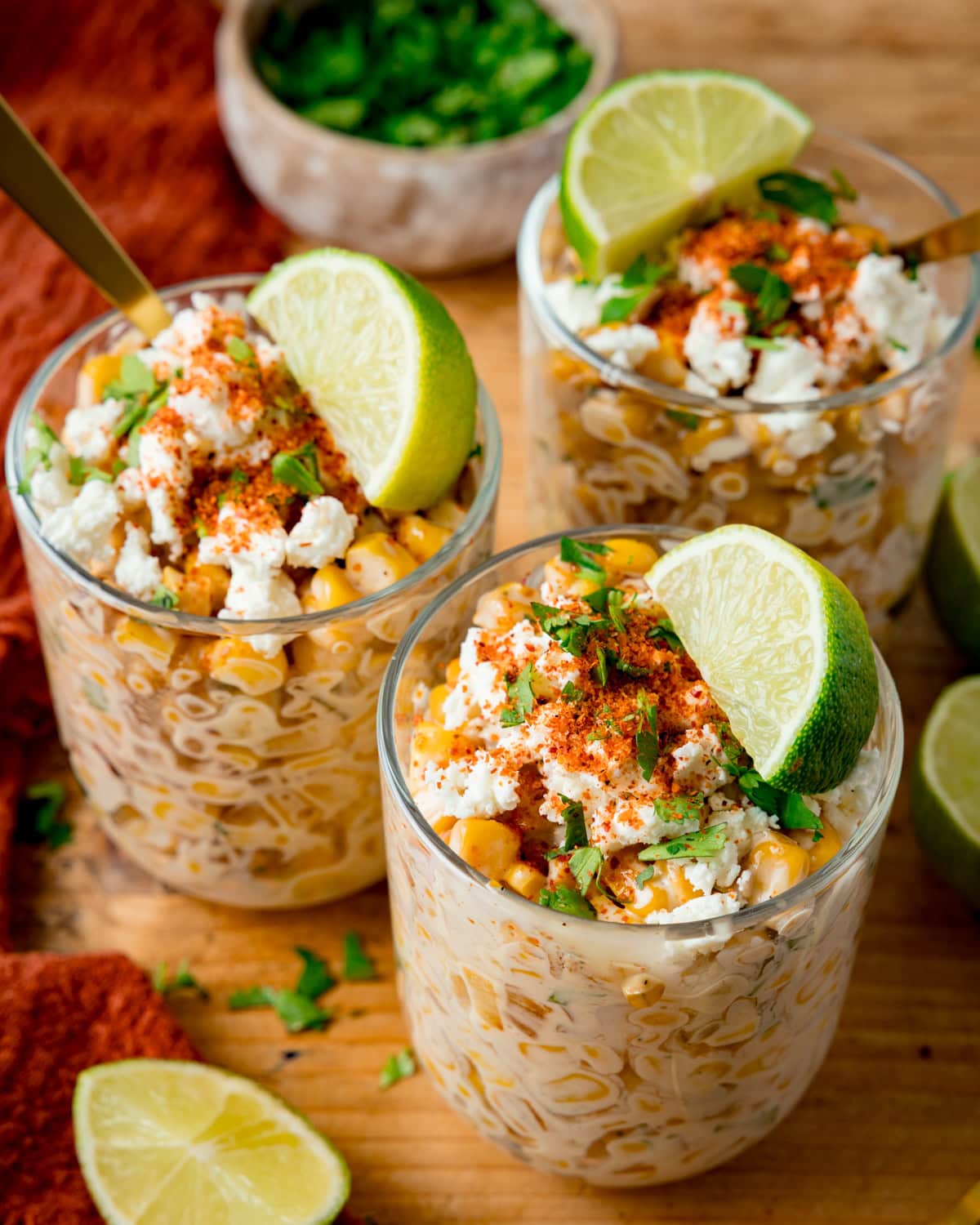 Three glasses filled with Mexican street corn salad—Esquites—topped with crumbled cheese, Tajin powder, cilantro, and a lime wedge on a wooden surface.