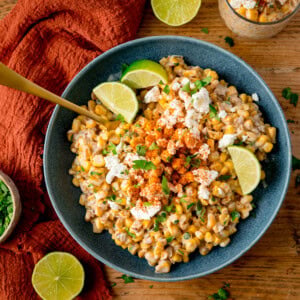 A blue bowl filled with Mexican street corn salad, also called Corn Esquites, topped with crumbled cheese, Tajin powder, and lime wedges, with a gold spoon in the corn. The bowl is on a wooden surface, next to a terracotta napkin and lime pieces.