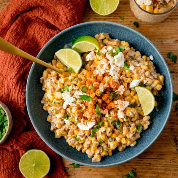 A blue bowl filled with Mexican street corn salad, also called Corn Esquites, topped with crumbled cheese, Tajin powder, and lime wedges, with a gold spoon in the corn. The bowl is on a wooden surface, next to a terracotta napkin and lime pieces.
