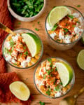 Three glasses filled with corn esquites, a Mexican street corn salad, topped with crumbled cheese, chilli powder, chopped coriander, and lime wedges on a wooden table.