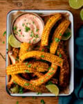 Overhead image of corn ribs served in a tray with lime wedges, chopped herbs, and a bowl of creamy dipping sauce on a wooden table.