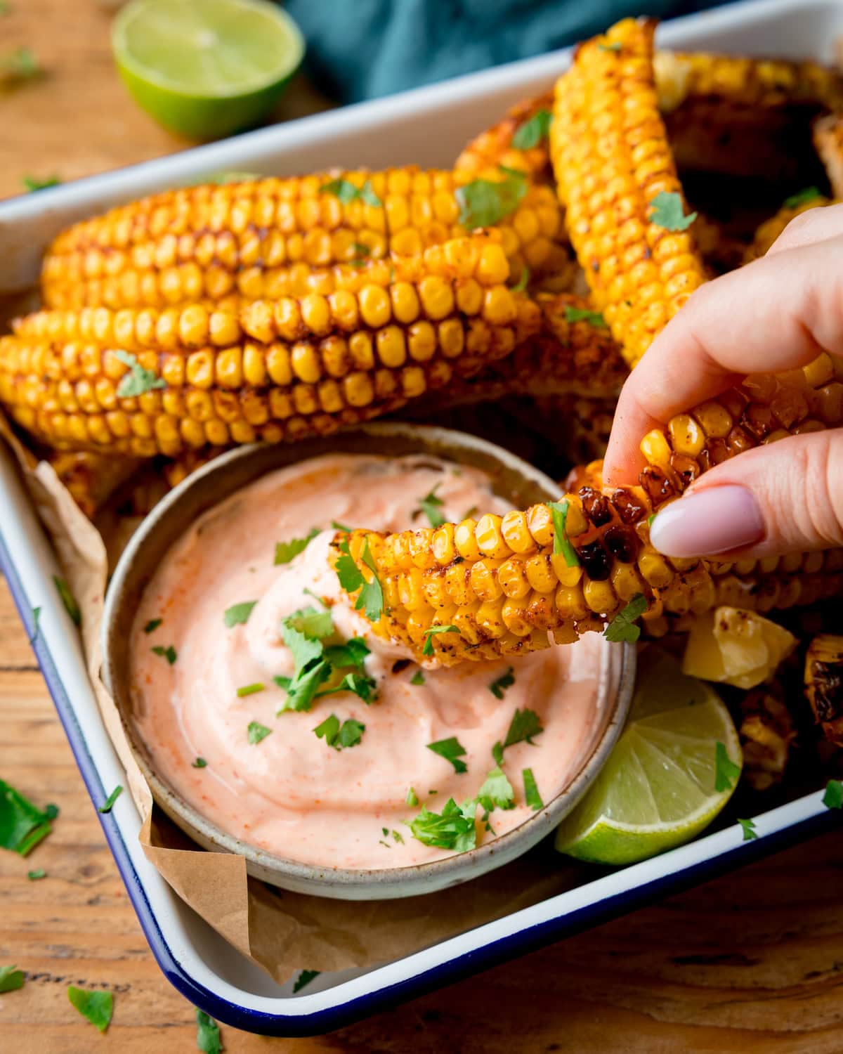 A hand dips a piece of grilled corn on the cob into a bowl of creamy sauce, garnished with chopped herbs. More corn ribs are piled up on a tray, which sits on a wooden surface.