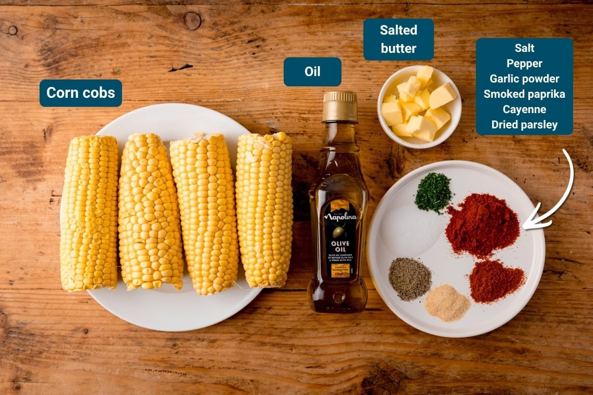 Four corn cobs, olive oil, salted butter, and a plate of spices on a wooden surface. All ingredients labelled - for corn ribs recipe.