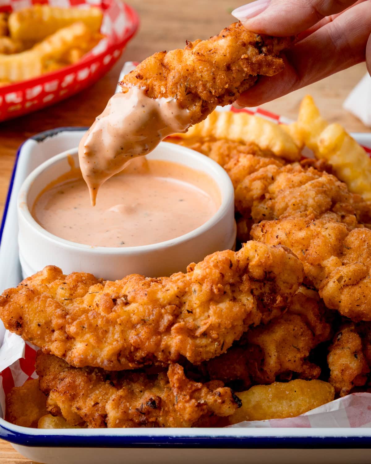 A hand dips a crispy fried chicken tender into creamy Cane's sauce, with more tenders and crinkle-cut fries in a tray nearby.