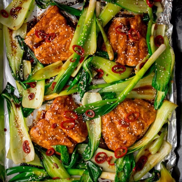 Salmon fillets and pak choi topped with sliced red chilli and sesame seeds, arranged on a foil-lined baking tray.