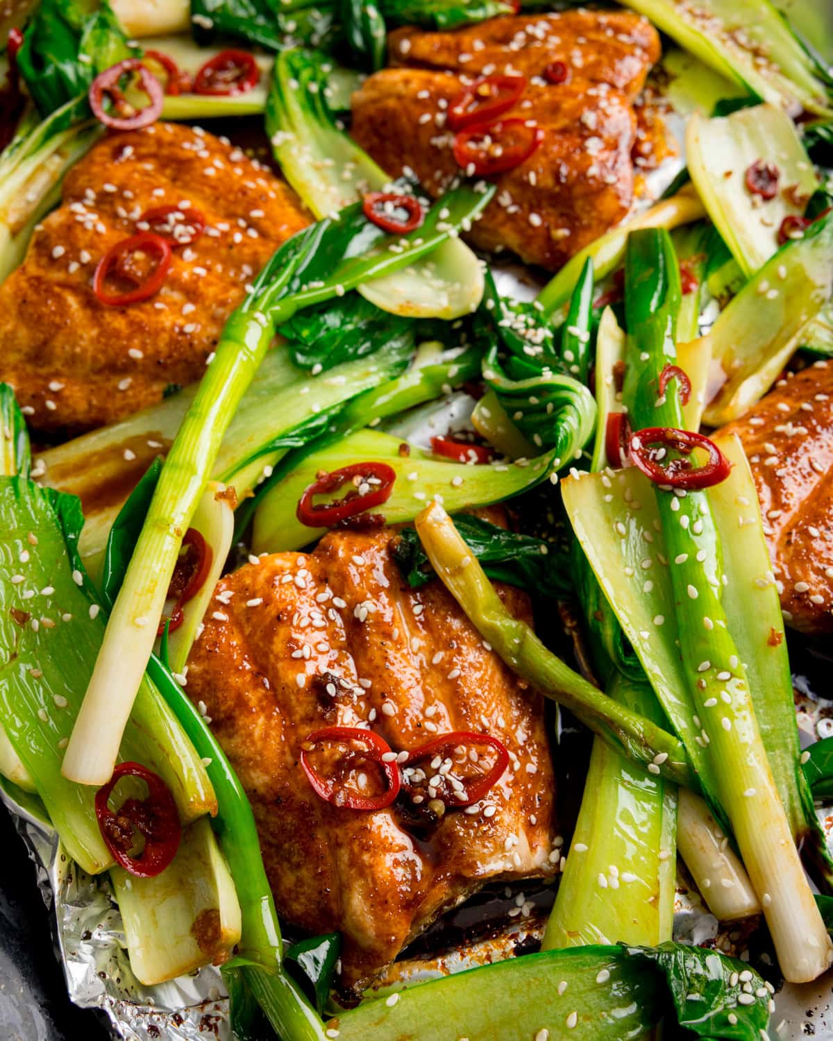Soy Glazed Salmon fillets topped with sesame seeds and sliced red chilli, served with cooked pak choi and spring onions on a foil-lined tray.