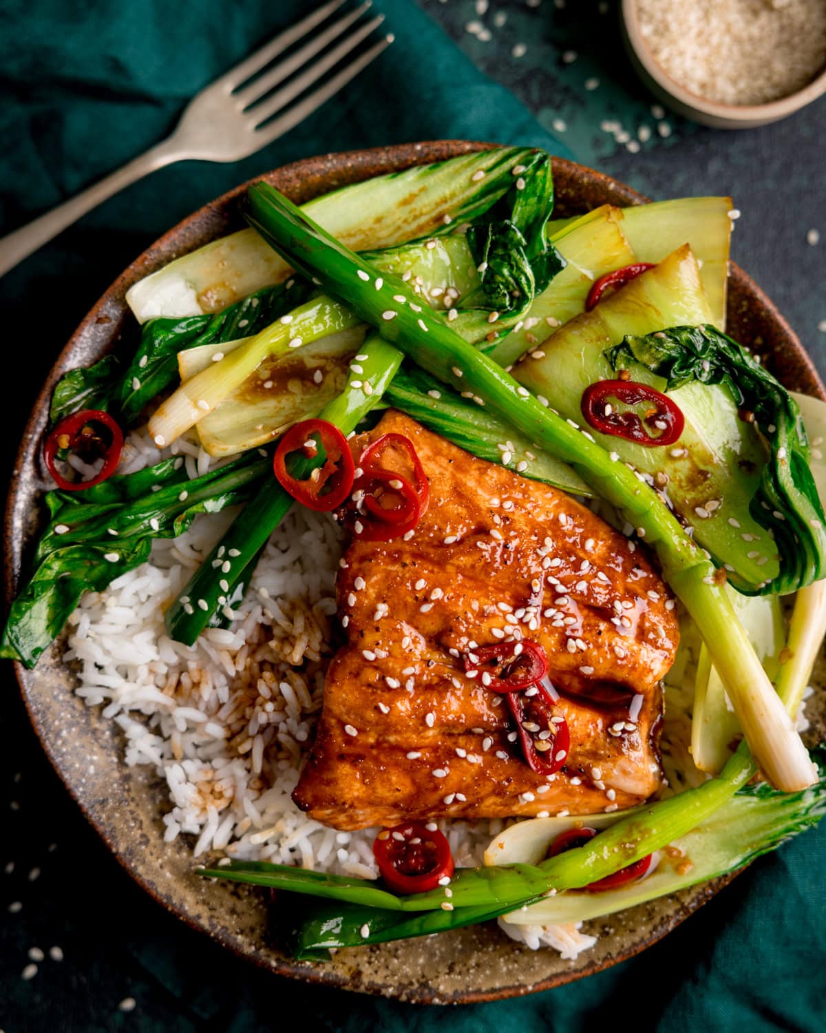 A bowl of white rice topped with glazed tofu, sautéed bok choy, spring onions, red chilli slices, and sprinkled sesame seeds.