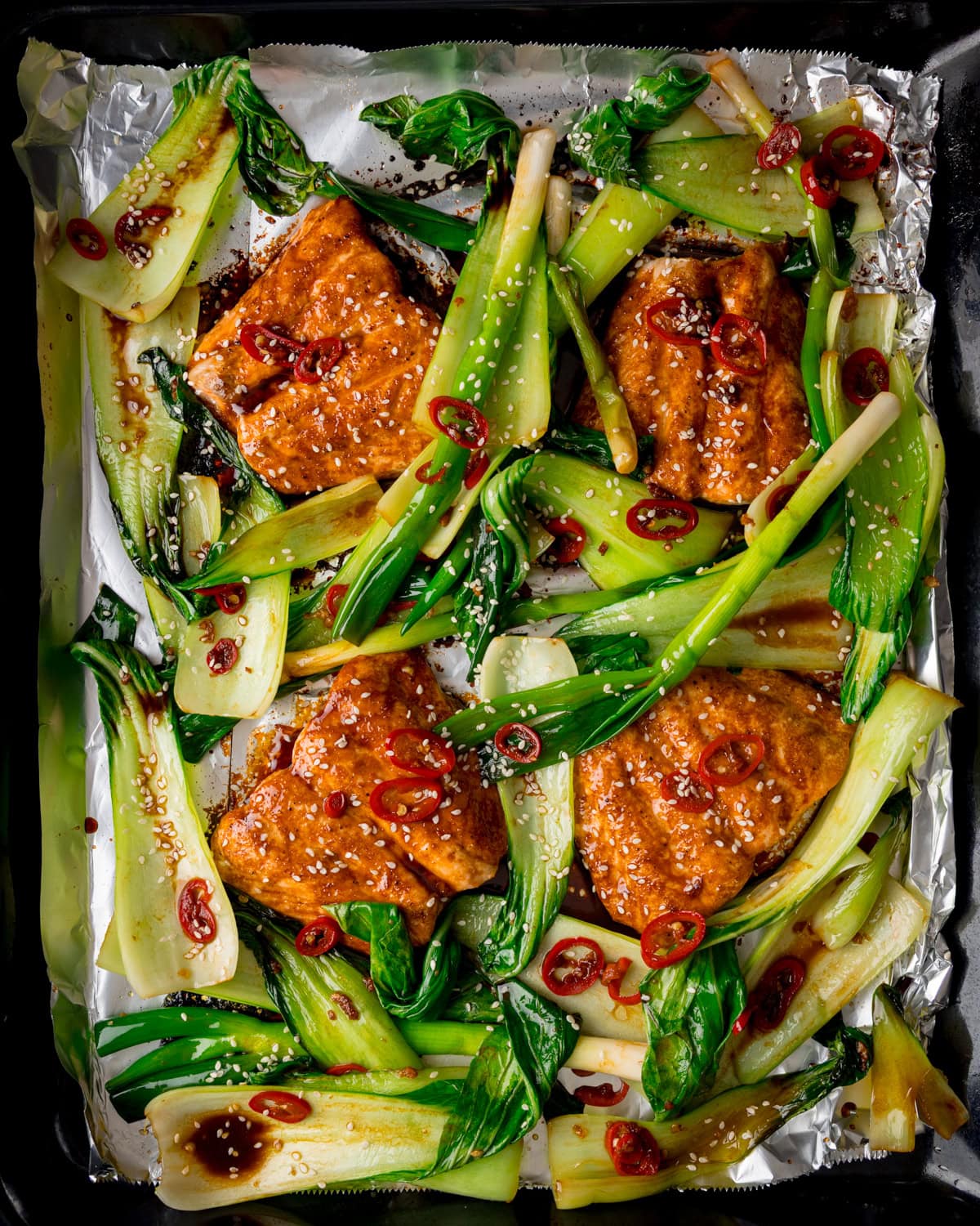 Soy Glazed Salmon fillets and stir fried pak choi on a foil-lined tray, garnished with sliced red chilli, sesame seeds, and a brown sauce.