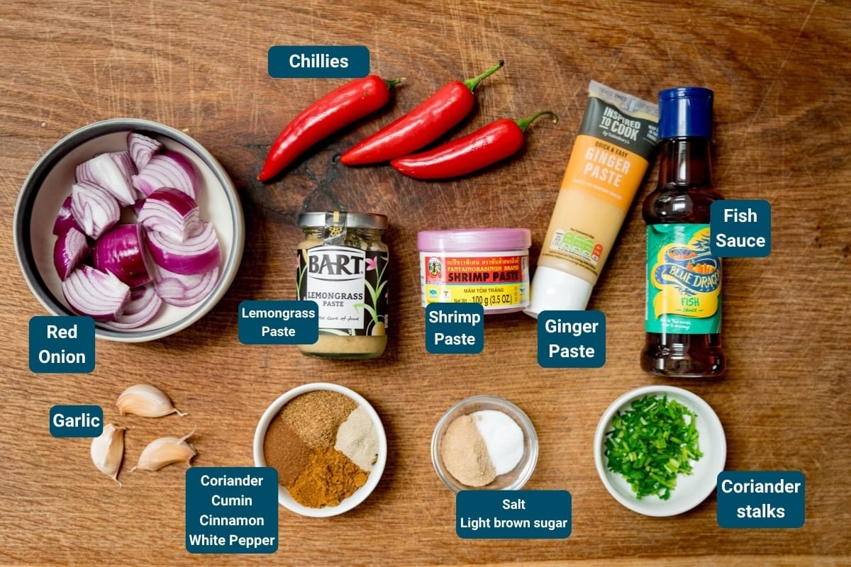 Ingredients for Massaman Curry Paste on a wooden surface, including red onion, garlic, chillies, ginger paste, shrimp paste, lemongrass paste, fish sauce, coriander stalks and spices. All ingredients are labelled.