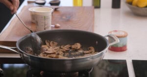 A person stirs sliced mushrooms, onions and garlic in a frying pan on a hob, with kitchen ingredients and utensils visible in the background.
