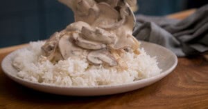 A plate of white rice topped with a creamy mushroom stroganoff being served, set on a wooden table with a grey napkin in the background.