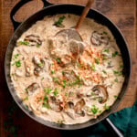 A frying pan filled with creamy mushroom stroganoff, garnished with chopped parsley, and paprika, with a wooden spoon inside.