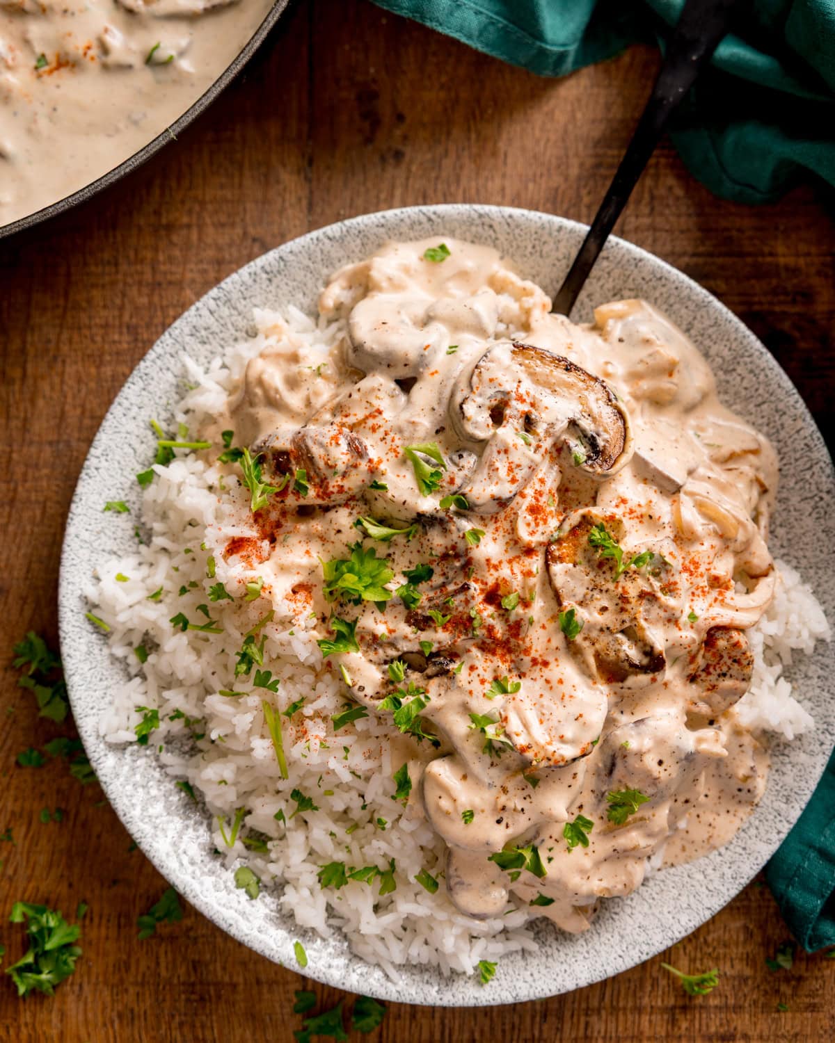 A plate of white rice topped with creamy mushroom stroganoff, garnished with chopped parsley and paprika, on a wooden table.