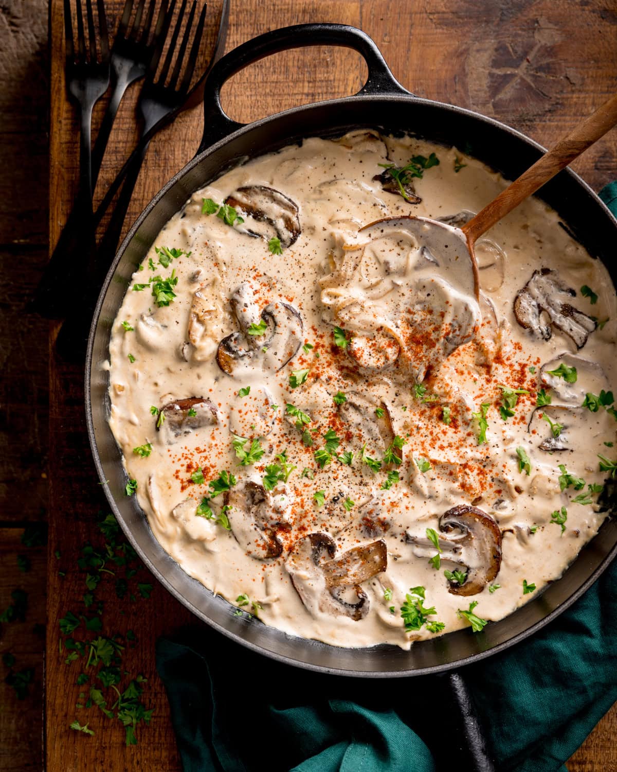 A frying pan filled with creamy mushroom stroganoff garnished with chopped parsley and paprika, with a wooden spoon inside. Four forks rest on the table nearby.