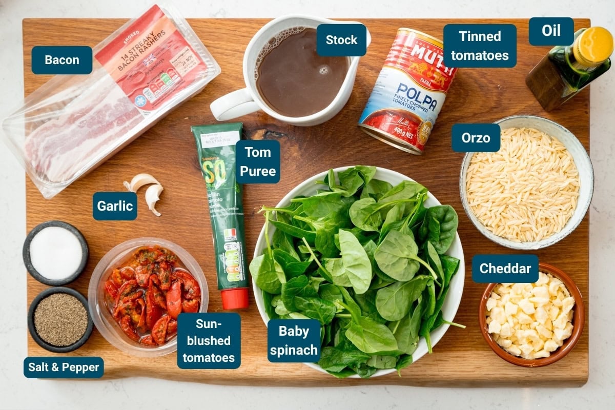 A wooden board displays labelled ingredients for a recipe: bacon, garlic, sun-blushed tomatoes, salt and pepper, baby spinach, tomato purée, stock, tinned tomatoes, oil, orzo and cheddar.