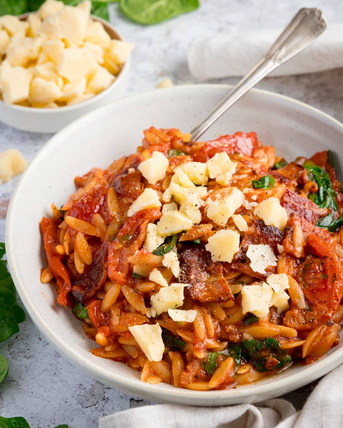 A bowl of tomato bacon orzo pasta in tomato sauce with spinach and sun-dried tomatoes, topped with chunks of cheese. A bowl of cheese sits in the background.