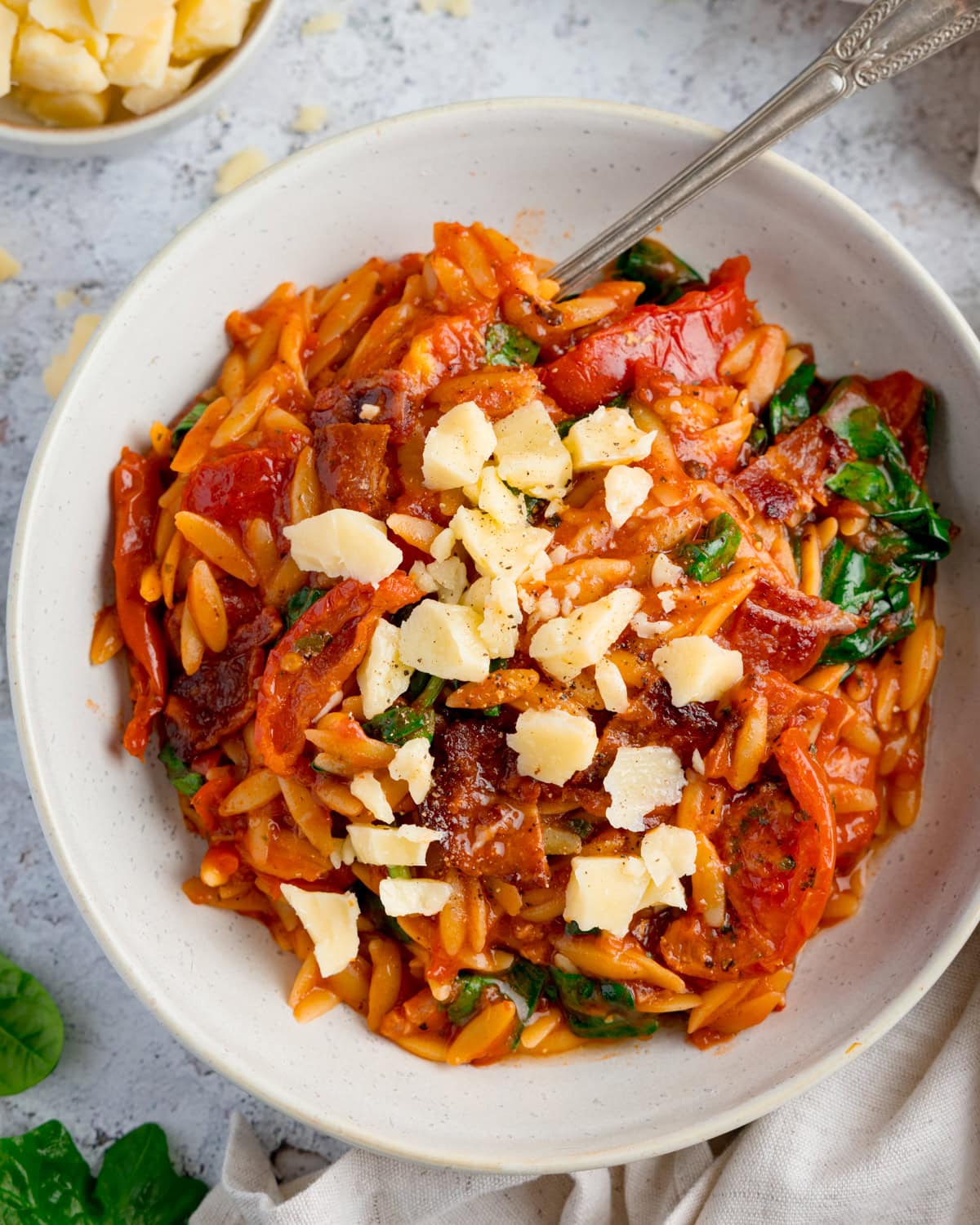 A bowl of orzo pasta in tomato sauce with spinach, sliced tomatoes, bacon, and topped with shaved cheese. A fork rests in the bowl.