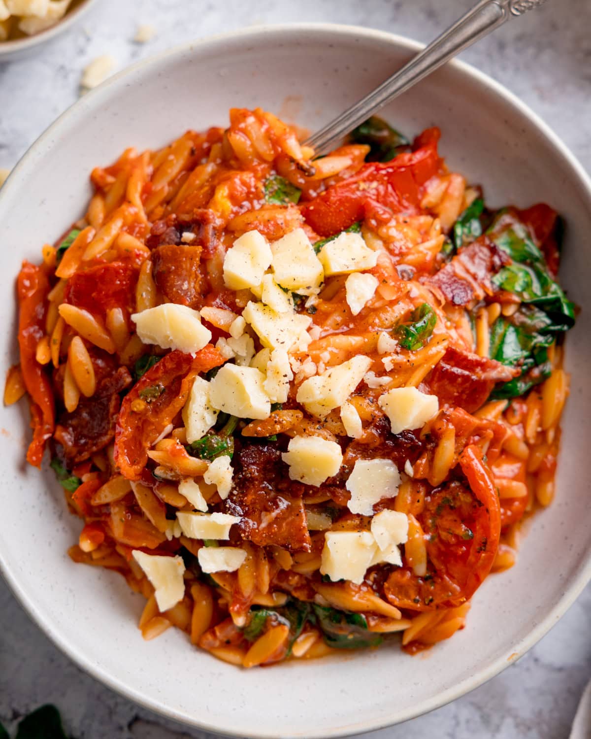 A bowl of tomato & bacon orzo pasta with spinach, sliced tomatoes, crispy bacon pieces, and cheese chunks on top, served with a fork.
