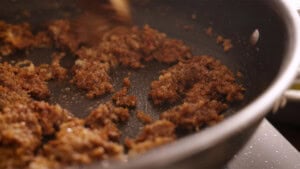 Minced onions, garlic and spices are being browned and cooked in a black frying pan on a hob, with some browning and texture visible.