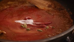 Tomato sauce simmering in a pan with cardamom pods, a pile of sugar, and a wooden spoon partially in view.