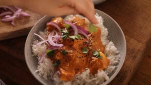 A hand garnishing a bowl of rice topped with creamy orange paneer curry, sliced red onions, coriander, and black seeds on a wooden table.