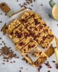 Peanut butter granola bars with oats and dried cranberries rest on a parchment-lined wooden board, next to scattered chocolate chips and dried cranberries and a cup of coffee.