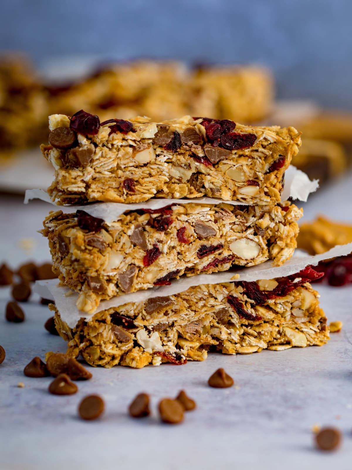 Three peanut butter granola bars with oats, nuts, chocolate chips and dried cranberries are stacked, separated by baking paper. Chocolate chips are scattered nearby.
