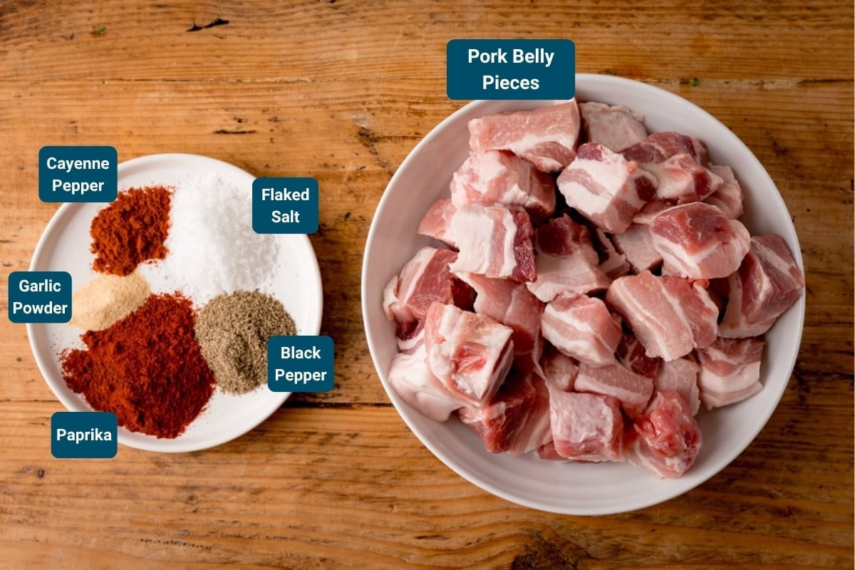 A bowl of raw pork belly pieces sits beside a small plate with paprika, garlic powder, cayenne pepper, black pepper, and flaked sea salt on a wooden surface.