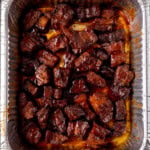 Overhead image of chunks of glazed Pork Belly Burnt Ends arranged in a single layer inside a disposable aluminium tray, coated in a dark, shiny sauce.