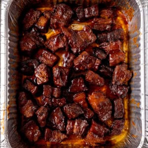 Overhead image of chunks of glazed Pork Belly Burnt Ends arranged in a single layer inside a disposable aluminium tray, coated in a dark, shiny sauce.