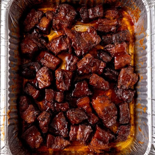 Overhead image of chunks of glazed Pork Belly Burnt Ends arranged in a single layer inside a disposable aluminium tray, coated in a dark, shiny sauce.